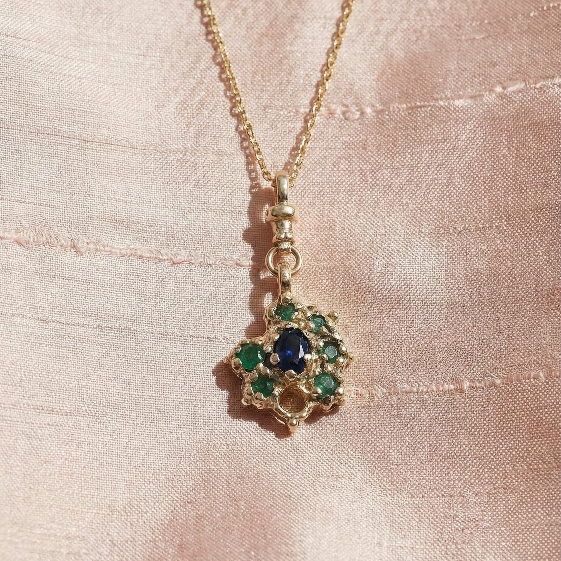 9ct Gold necklace with a pendant featuring emeralds and a central sapphire, on a pink silk background
