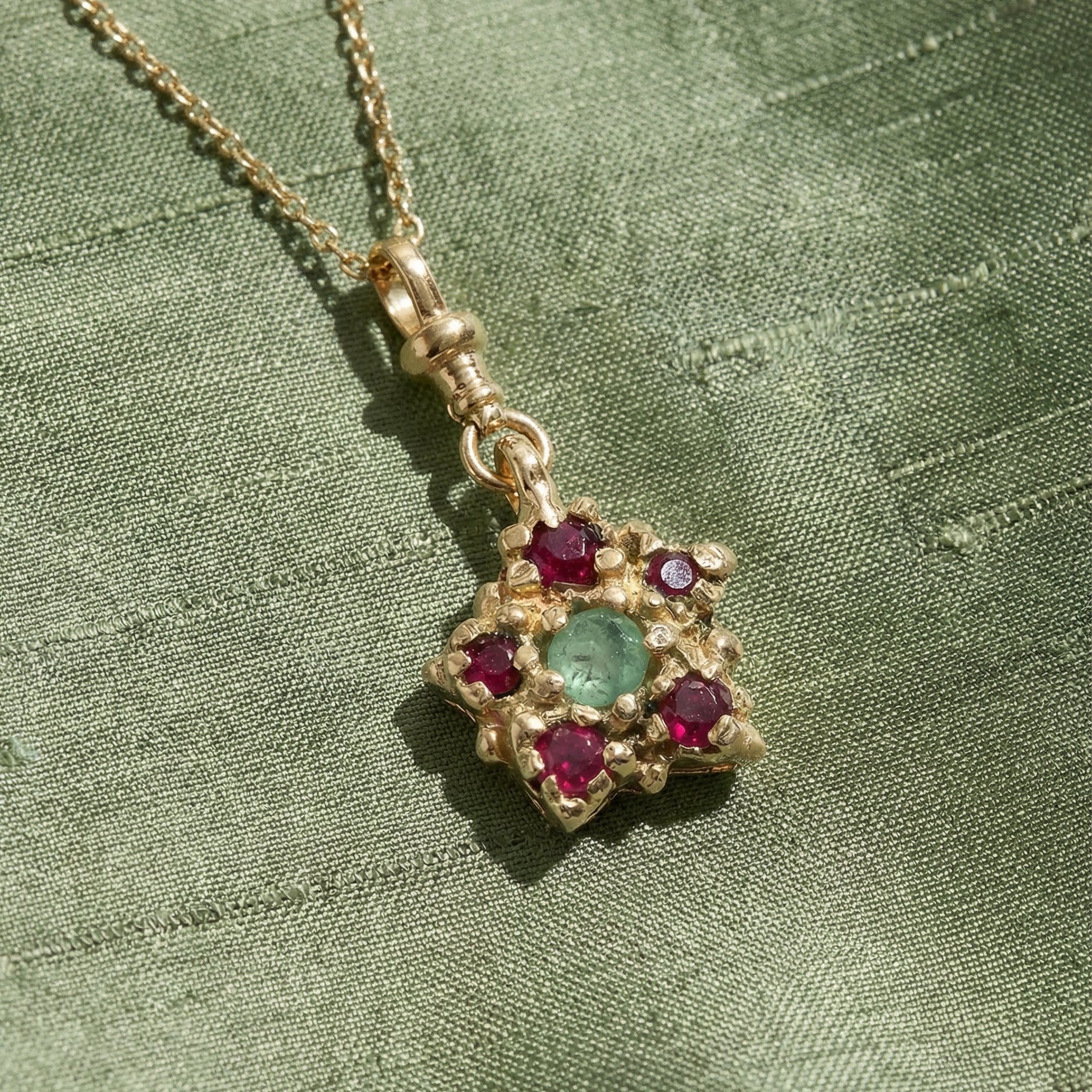 9ct yellow gold, victorian inspired, charm Necklace set with a central emerald and an inclusion, and surrounding rubies on a sage silk green fabric background
