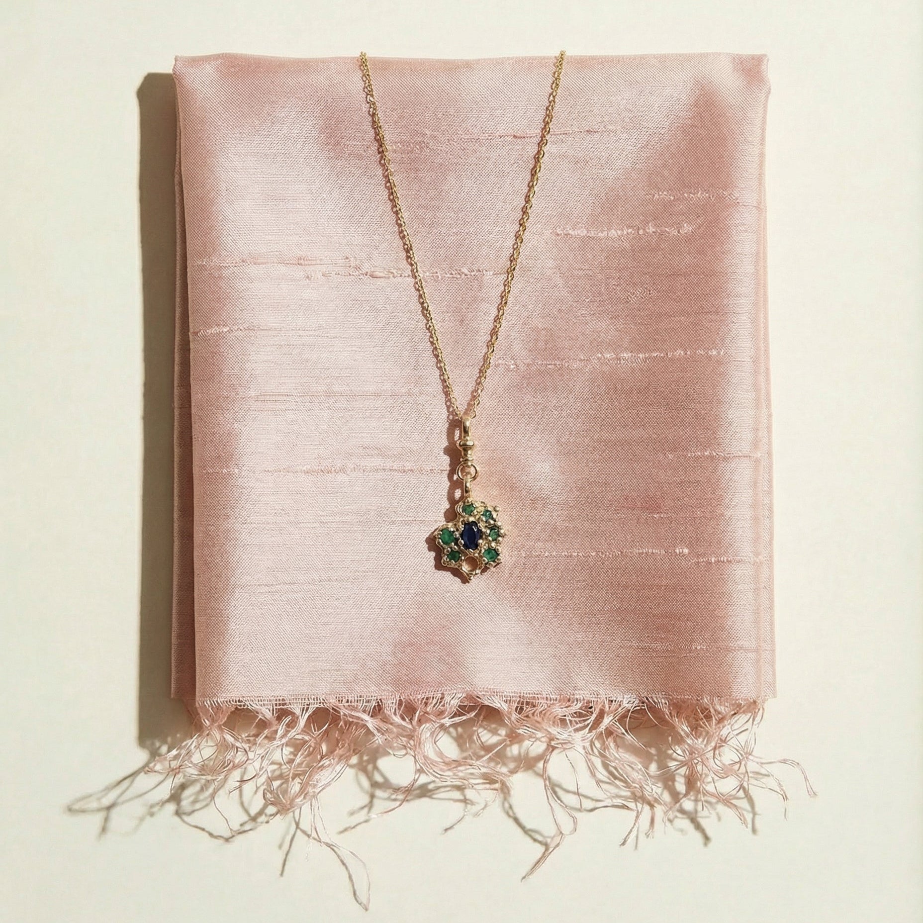9ct gold necklace with a emeralds and central sapphire pendant on a pink fabric background