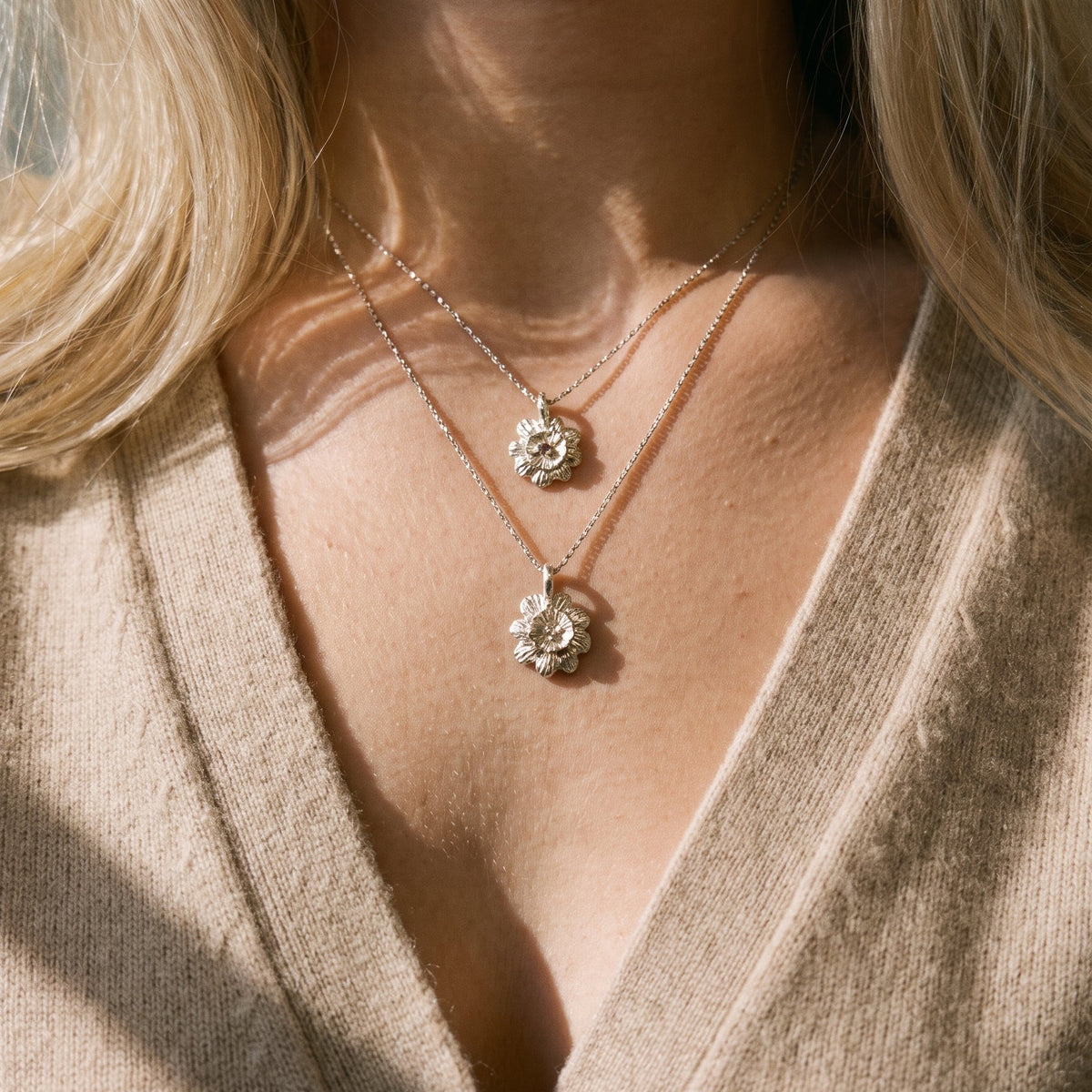 A close-up of a woman wearing two delicate silver necklaces with textured floral or starburst pendants over a beige knit top. The shot is lit by warm, direct sunlight casting shadows on her skin and clothing.