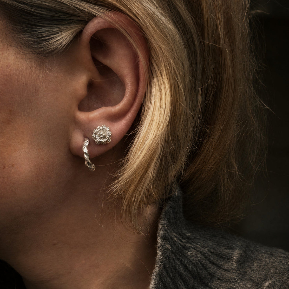A close-up of a person's ear wearing a small silver floral stud earring and a silver spiral hoop earring, with short blonde hair and a dark ribbed turtleneck sweater.