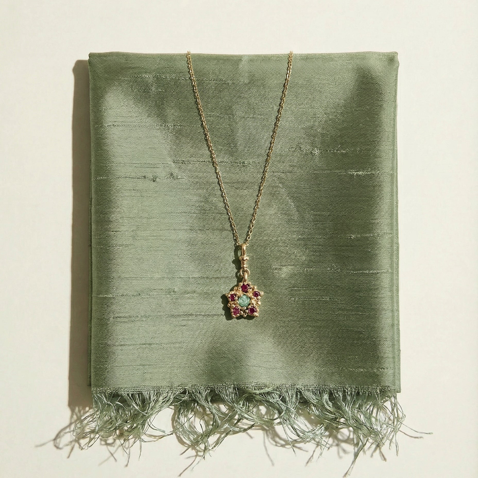9ct yellow gold, victorian inspired, charm Necklace set with a central emerald and an inclusion, and surrounding rubies on a sage silk green fabric background
