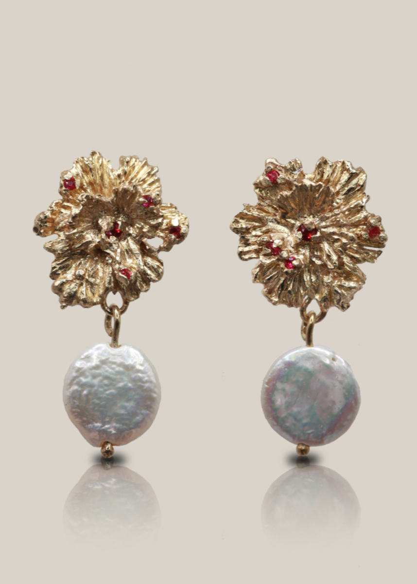 Pair of gold floral earrings with red gemstones and pearl-like bottoms on a light gray background