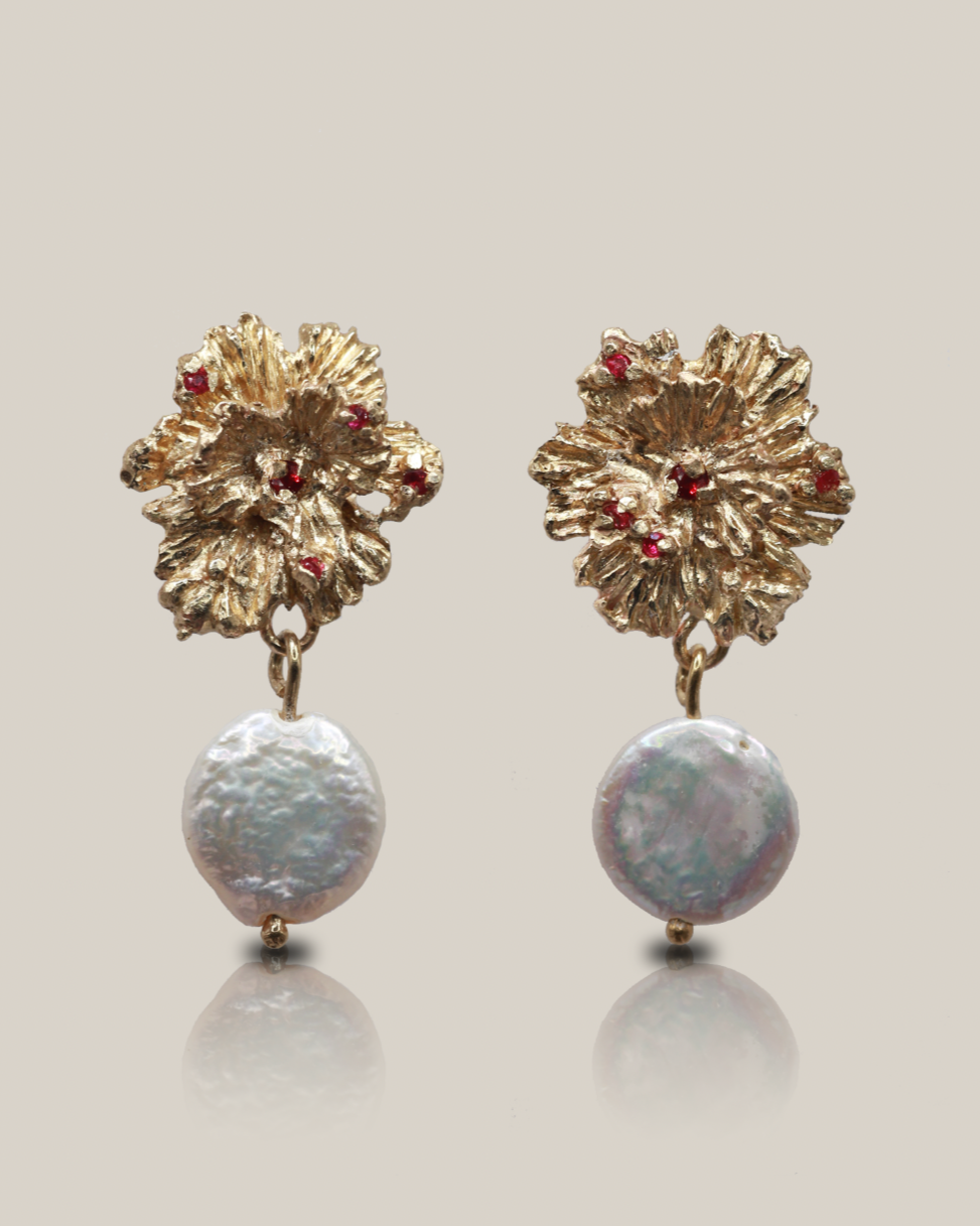 Pair of gold floral earrings with red gemstones and pearl-like bottoms on a light gray background