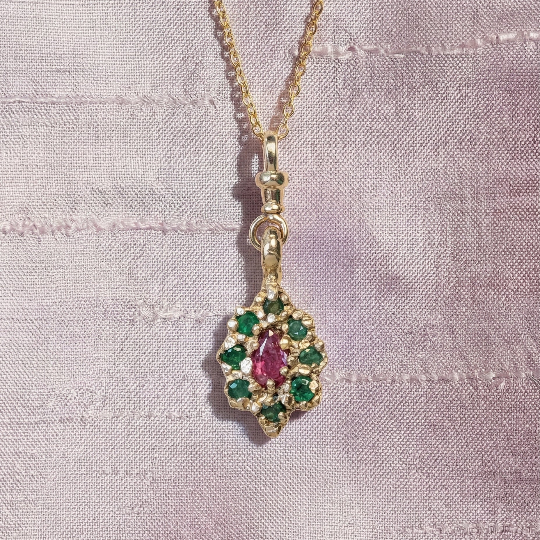 9ct Gold victorian inspired necklace with a pendant featuring emeralds and a central ruby gemstone, on a light purple fabric background